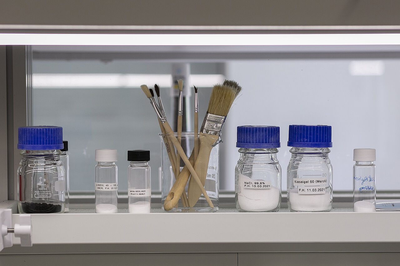Laborflaschen, gefüllt mit kristallinen Proben, und Laborwerkzeuge wie z.B. Pinsel.